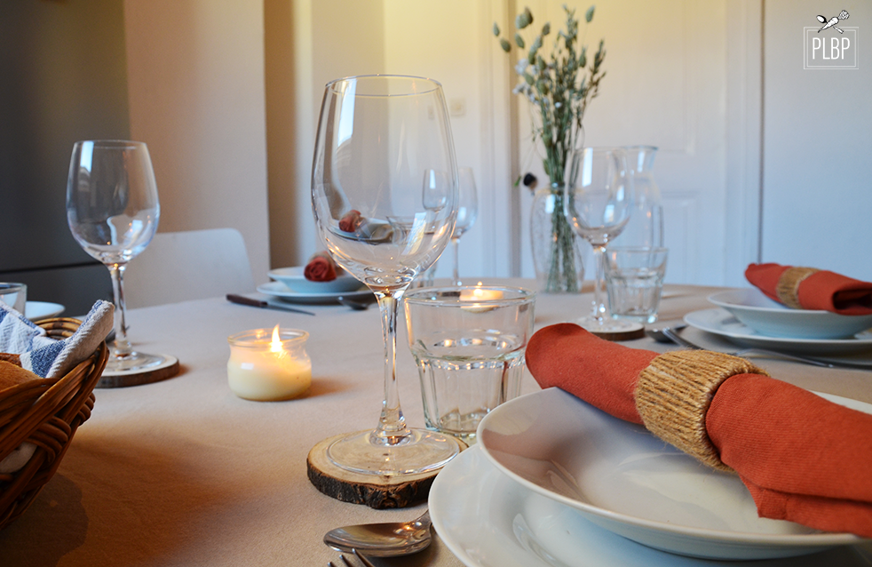 Photo d'une table pour 4 personnes vu de côté avec verres à vin et à eau, assiette et serviettes en tissus, couverts, fleurs séchées dans vase transparant et bougies.